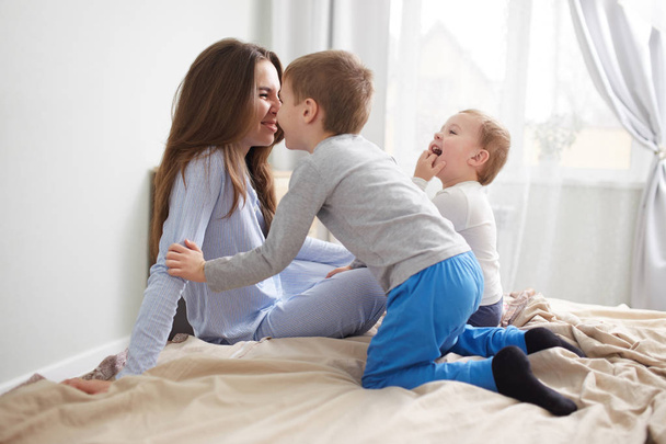 Young beautiful mother and her two little sons dressed in pajamas have fun on the bed in the light cozy bedroom - Fotó, kép