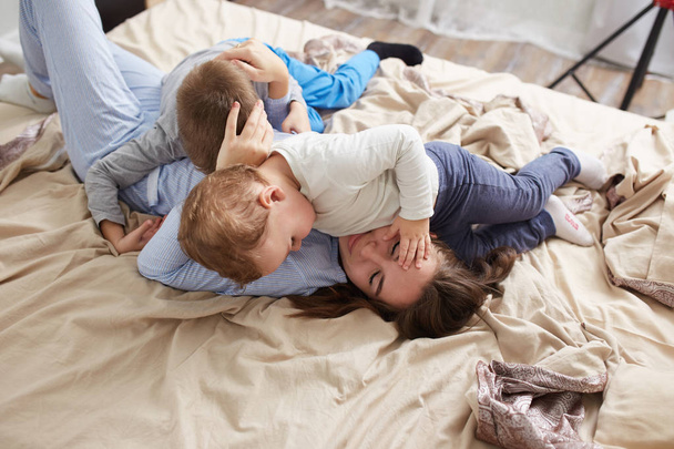 Beautiful young mother dressed in light blue pajama  and her two little sons have fun on the bed in the light cozy bedroom - Valokuva, kuva