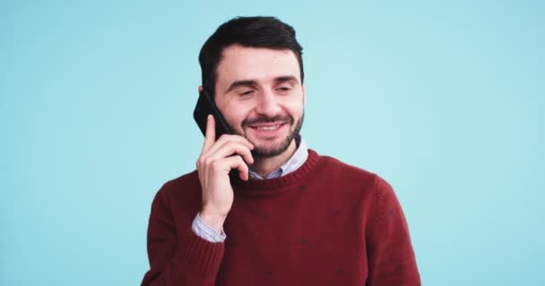 Smiling charismatic young man speaking on his phone in front of the camera in the studio with a blue background wall - Materiał filmowy, wideo