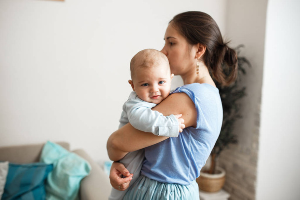 Young mother dressed in light blue t-shirt and skirt is holding her tiny son on her arms in the room at home - Foto, afbeelding