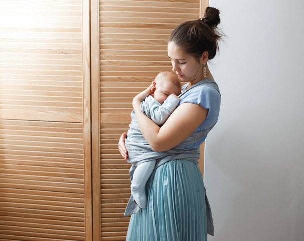 Young mother dressed in light blue t-shirt and skirt is holding her tiny son on her arms in the room next to the wooden doors - Zdjęcie, obraz