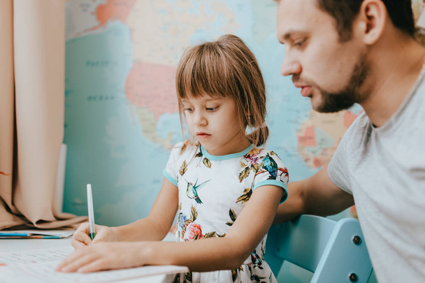 Father helps his little daughter to write at the desk in the room with the map on the wall - Fotó, kép