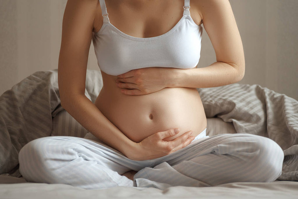 Primer plano de la mujer embarazada con dolor abdominal en camiseta blanca y pantalones sentados en la cama en casa sosteniendo animales a los lados
 - Foto, imagen