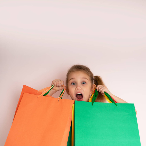 Ψώνια μικρό κορίτσι κρατώντας σακούλες για ψώνια κοιτάζοντας μέχρι το πλάι στο βάθος του φόντου στο χώρο αντιγραφής.. - Φωτογραφία, εικόνα