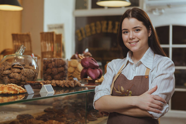 Charmante jonge vrouwelijke baker glimlachen naar de camera, trots te poseren in haar eigen bakkerij Cafe. Gelukkig vrouwelijke banketbakker genieten van werken bij haar bakkerij, kopie ruimte. Concept van het ondernemerschap - Foto, afbeelding