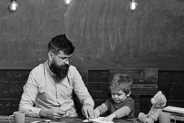 Teacher and little kid sitting at desk with books and teddy bear. Schoolboy playing with copybook. Strict teacher in glasses in front of green board - Valokuva, kuva