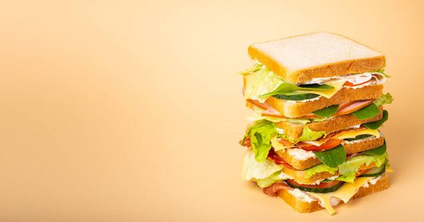 Close up of tall whole tasty sandwich with cheese, ham, prosciutto, fresh lettuce, tomatoes, cucumbers on pastel yellow background. Healthy gourmet sandwich for breakfast or lunch. Space for text  - Photo, image