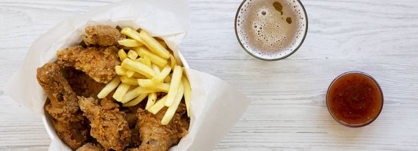 Tasty fastfood: fried chicken drumsticks, spicy wings, French fries and chicken tenders in paper box, sauce and cold beer on a white wooden surface, overhead view. Flat lay, top view, from above.  - Fotoğraf, Görsel