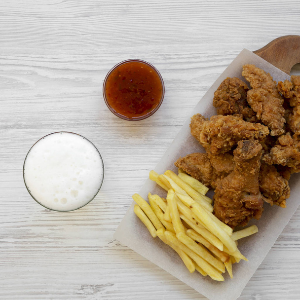 Tasty fastfood: fried chicken drumsticks, spicy wings, French fries and chicken fingers with sour-sweet sauce and cold beer over white wooden surface, top view. Flat lay, overhead, from above.  - Photo, Image