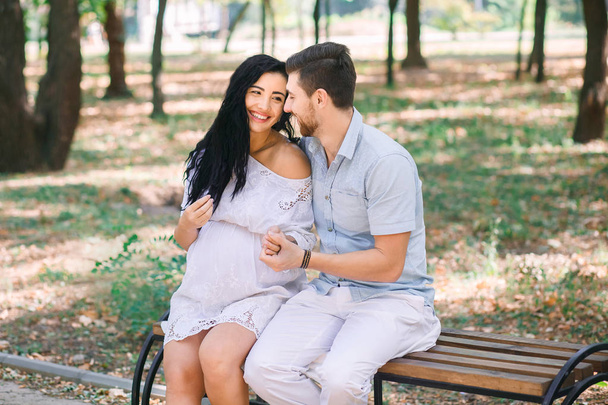Ein junger Mann und seine schwangere Frau sitzen auf einer Bank, vor dem Hintergrund von Grün und Bäumen. in Erwartung eines Kindes. erste Schwangerschaft ich liebe Kinder. - Foto, Bild