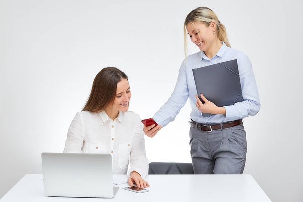 Blonde caucasian female manager shows something on the cell phone to her brunette female colleague with a smile in the office with the white background. Girls laugh together. - Photo, Image