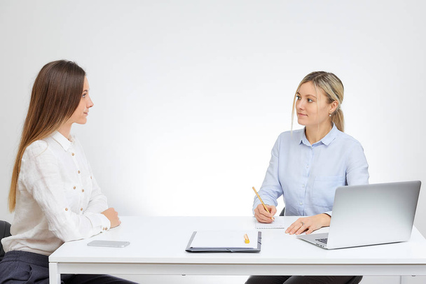 Blonde caucasian female boss in a blue shirt interviews new brunette female employee sits by the desk in the office with the white background. - Fotografie, Obrázek