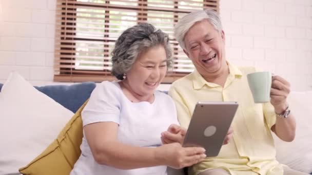 アジアの高齢者のカップル タブレットを使用して、自宅のリビング ルームでコーヒーを飲みながら、カップルは家でゆっくりとソファの上に横たわっている間愛の瞬間をお楽しみください。ホーム コンセプトで時間ライフ スタイル シニア家族を楽しんでください。. - 映像、動画
