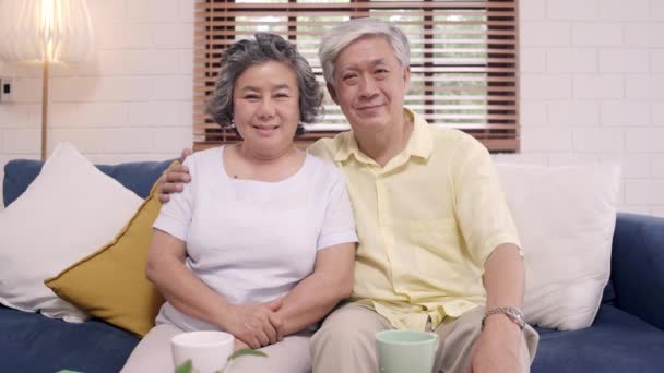 感じて幸せ笑顔としながらカメラをアジアの老夫婦は、自宅のリビング ルームでソファの上リラックスします。時間ホーム コンセプト シニア家族ライフ スタイルを楽しんでいます。カメラ目線の肖像画. - 映像、動画