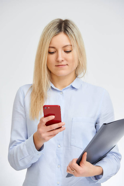 Молодая милая блондинка-кавказка смотрит на свой красный сотовый телефон, держа папку с документами в офисе на белом фоне
. - Фото, изображение