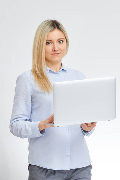 Young cute blonde caucasian woman with smile stands sideways holding her aluminum notebook computer  in the office with a white background. - Photo, Image