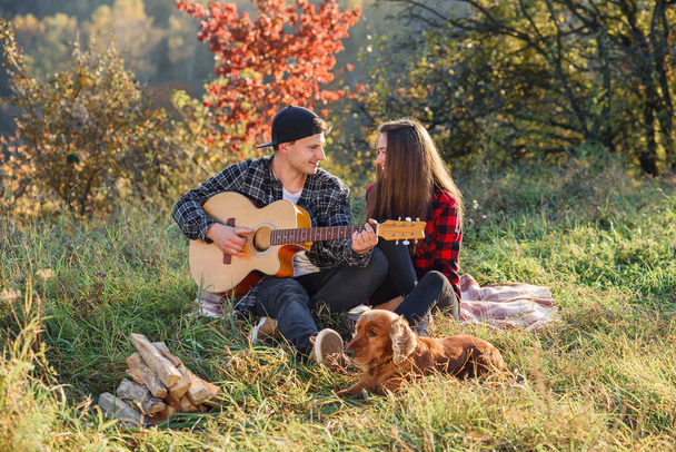 Happy caucasian couple with guitar and their dog having rest on picnic in the park on the lawn. - Foto, afbeelding
