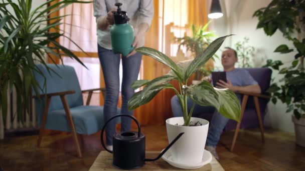 jonge vrouw die het sproeien van water op plant in moderne huis man leest uit tabletwater op plant met sprinkler - Video