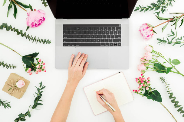 Woman work with laptop. Workspace with female hands, laptop, notebook and pink flowers on white background. Top view. Flat lay. - Photo, Image