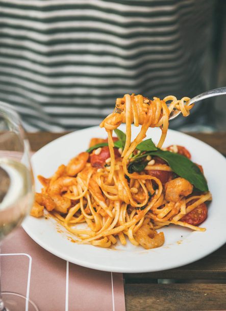 Italian dinner at bistrot with spaghetti pasta with shrimps and glass of white wine. Man eating pasta with fork. Delicious Italian cuisine concept - Photo, Image