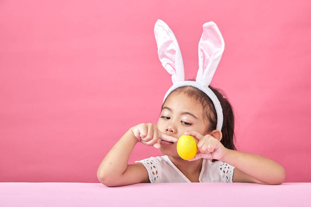 Щаслива азіатська маленька дівчинка, яка носить вуха кролика на Великдень. Дівчина тримає пофарбовані яйця, рожевий мінімальний фон
 - Фото, зображення