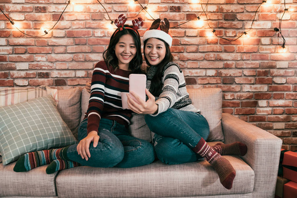 Happy female friends make selfie on Christmas Eve in Santa Claus sweaters and reindeer hat playing at home. Enjoying holidays Merry Christmas and Happy New year. girls using cellphone take photo - Foto, Bild