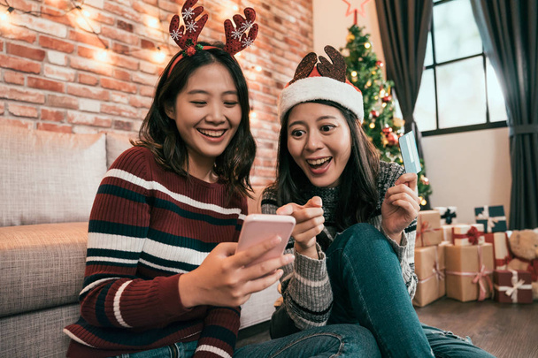 tourists ready for xmas new year holidays travel booking hotel online with credit card and mobile phone at home. young girls sitting on floor christmas tree. asian ladies shopping on boxing day sale - Foto, Bild