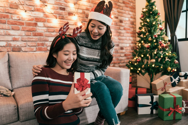 Two pretty sisters celebrating Christmas together sitting near New Year tree and opening holiday presents. Best friends hugs and having great time at home opening gift box smiling. girls in santa hat - 写真・画像