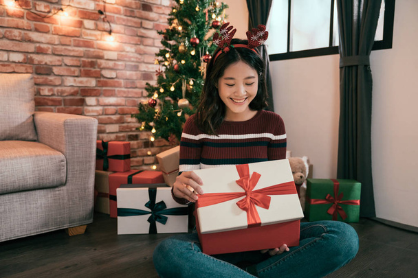 happy young woman in reindeer hat opening gift box shiny next to golden beautiful christmas tree with lights and presents in festive room. happy winter holiday atmospheric moments. seasons greetings - Photo, Image