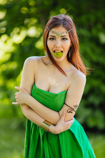 Portrait of a beautiful young asian woman in green dress and diadema on head holding slice of kiwi in mouth - Fotografie, Obrázek
