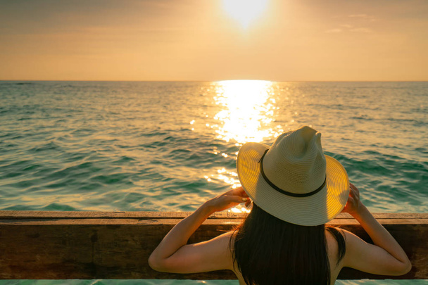 Back view of black long hair sexy Asian woman with straw hat relaxing and enjoying holiday at tropical paradise beach and watch the sunset sky. Summer vacation. Holiday travel alone. Summer vibes.  - Photo, Image