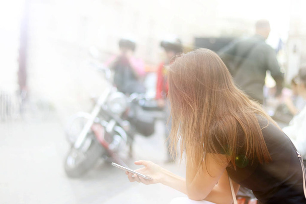 Een jong meisje is op zoek naar een smartphone die houdt. Een vrouw zit op een bankje in het centrum van een oude stad tegen een motorfiets en voorbijgangers in een wazig vervagen. Levensstijl en vrije tijd. - Foto, afbeelding