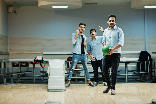 South asian man in jeans shirt standing at bowling alley with ball on hands. Guy is preparing for a throw. Friends support him loudly. - Foto, Bild