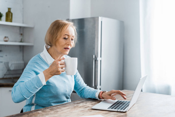 Seniorin sitzt am Tisch, trinkt Kaffee und benutzt Laptop in Küche mit Kopierraum - Foto, Bild