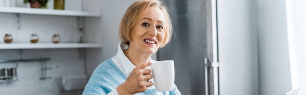 χαμογελώντας ανώτερων γυναίκα κρατώντας το φλιτζάνι του καφέ και κοιτάζοντας κάμερα στο σπίτι - Φωτογραφία, εικόνα