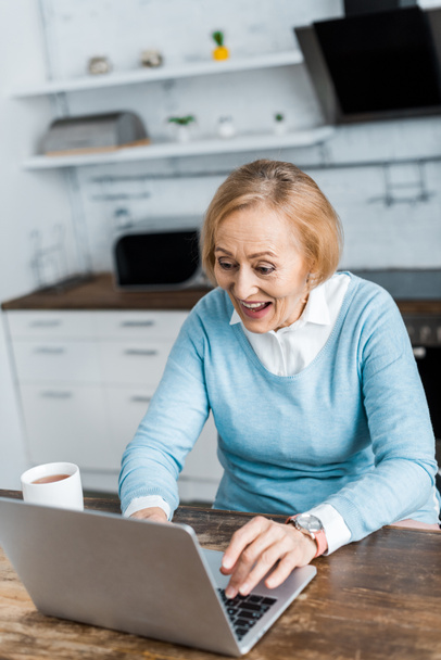 excited senior woman sitting at table with cup of tea and using laptop in kitchen - Photo, Image