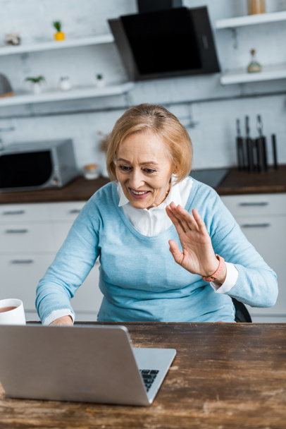 souriant femme âgée assise à la table et agitant la main tout en ayant appel vidéo sur ordinateur portable dans la cuisine
 - Photo, image