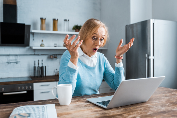 zaskoczony starszy kobieta gestem rękami, za pomocą laptopa i konieczności rozmowy wideo w kuchni - Zdjęcie, obraz