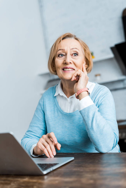 glimlachend peinzende senior vrouw zittend aan tafel met laptop en stutten kin thuis  - Foto, afbeelding