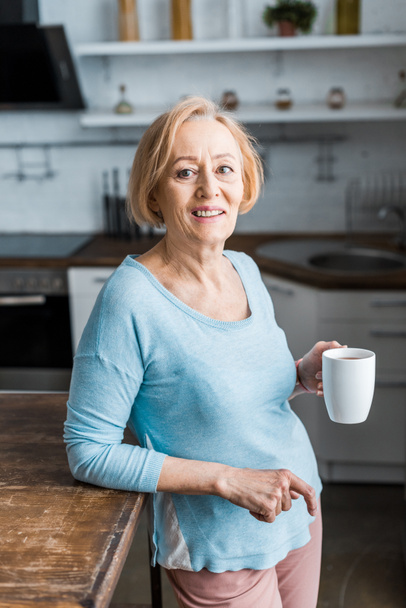 smiling senior woman with cup of coffee looking at camera in kitchen - Foto, afbeelding