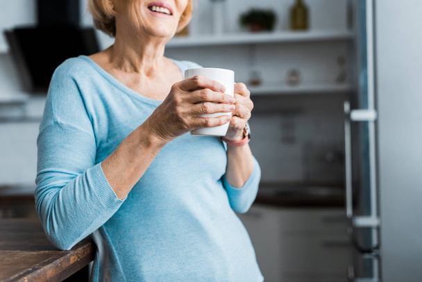 vista recortada de la mujer mayor sonriente sosteniendo taza de café en casa con espacio para copiar
 - Foto, Imagen