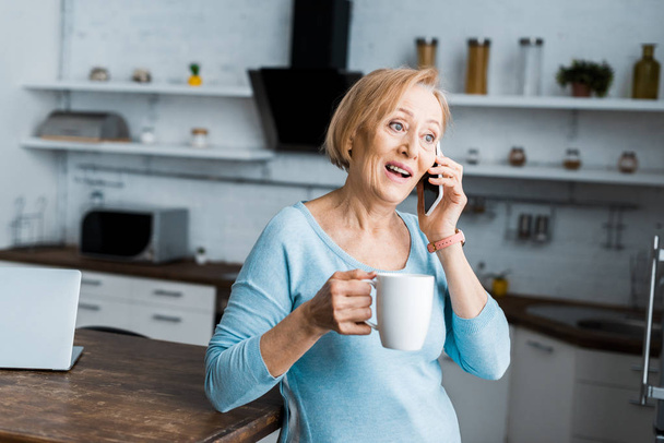überraschte Seniorin mit Tasse Kaffee im Smartphone-Gespräch zu Hause - Foto, Bild