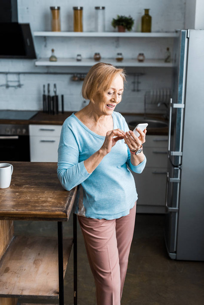 hymyilevä vanhempi nainen käyttää älypuhelinta keittiössä
  - Valokuva, kuva