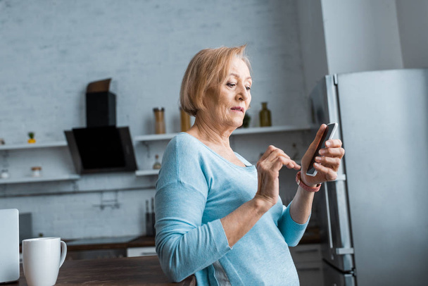 Senior vrouw met behulp van smartphone in keuken met kopie ruimte - Foto, afbeelding