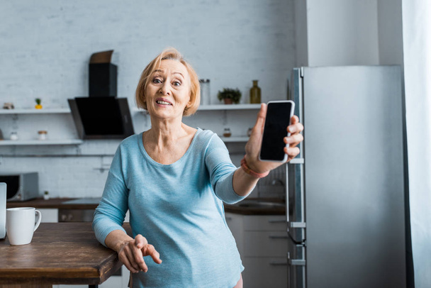 χαμογελώντας ανώτερων γυναίκα κοιτάζοντας φωτογραφική μηχανή και παρουσιάζουν smartphone με κενή οθόνη στο σπίτι - Φωτογραφία, εικόνα