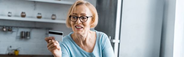 Lächelnde Seniorin mit Brille und Kreditkarte zu Hause mit Kopierplatz - Foto, Bild