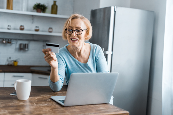 sorrindo mulher sênior segurando cartão de crédito e usando laptop ao fazer compras on-line em casa
 - Foto, Imagem