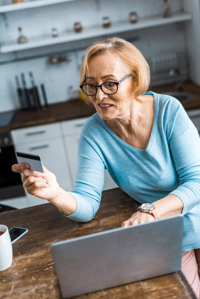 selective focus of senior woman holding credit card and using laptop while doing online shopping at home - Φωτογραφία, εικόνα