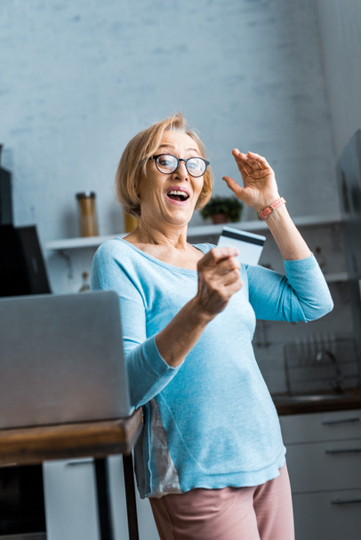 sorprendida mujer mayor en gafas con tarjeta de crédito cerca de la computadora portátil en casa
 - Foto, Imagen