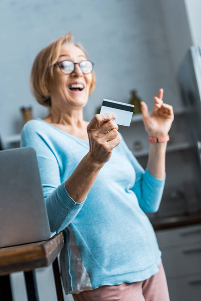 aufgeregte Seniorin mit Brille hält Kreditkarte in der Nähe von Laptop zu Hause - Foto, Bild